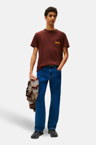 T-shirt brun à manches courtes de TOMMY JEANS, avec poche poitrine et porté avec des jeans bleus et des baskets noires.