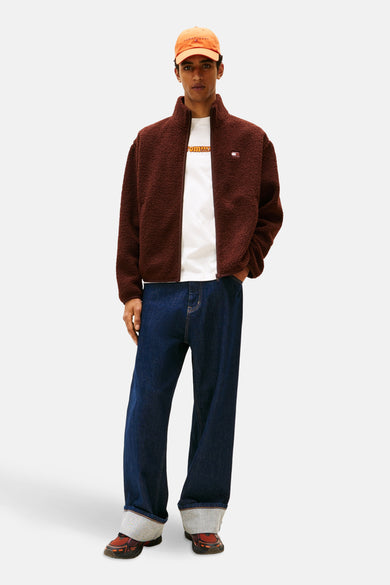 Veste brune de TOMMY JEANS avec fermeture éclair, portée sur un T-shirt blanc et des jeans bleu marine, avec un chapeau orange.