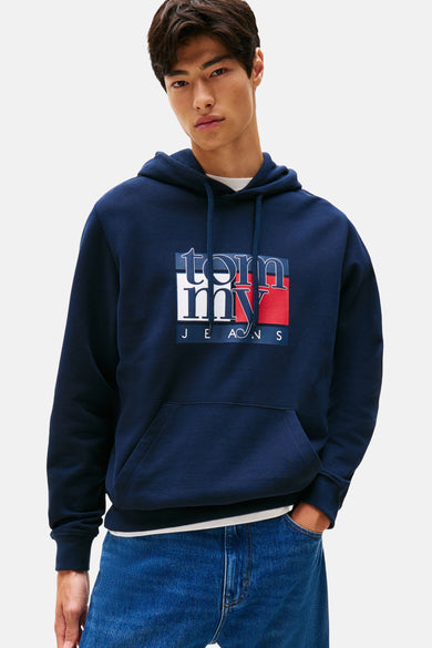 Pull-over bleu avec capuche de TOMMY JEANS, avec logo sur l'avant, porté avec un T-shirt blanc et des jeans bleus.