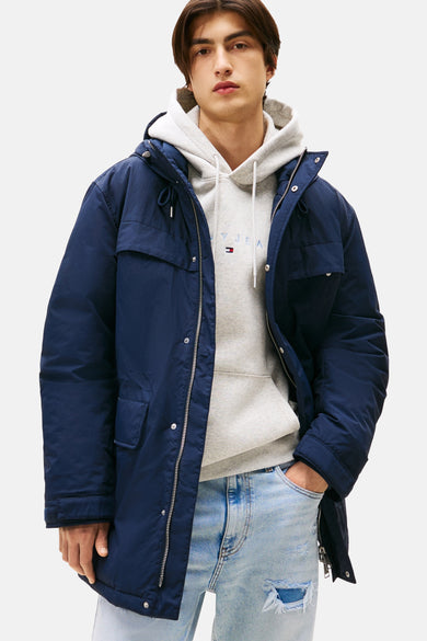 Manteau long bleu de TOMMY JEANS, avec capuche et porté sur un hoodie gris et des jeans déchirés.