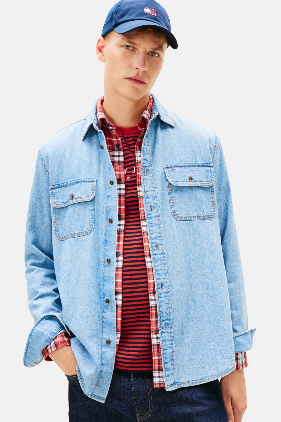 Man met een blauwe Tommy Jeans pet, denim overhemd, rood geruit overhemd en gestreepte trui.