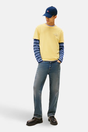 Man in gele T-shirt met blauwgestreepte longsleeve, jeans en blauwe pet.