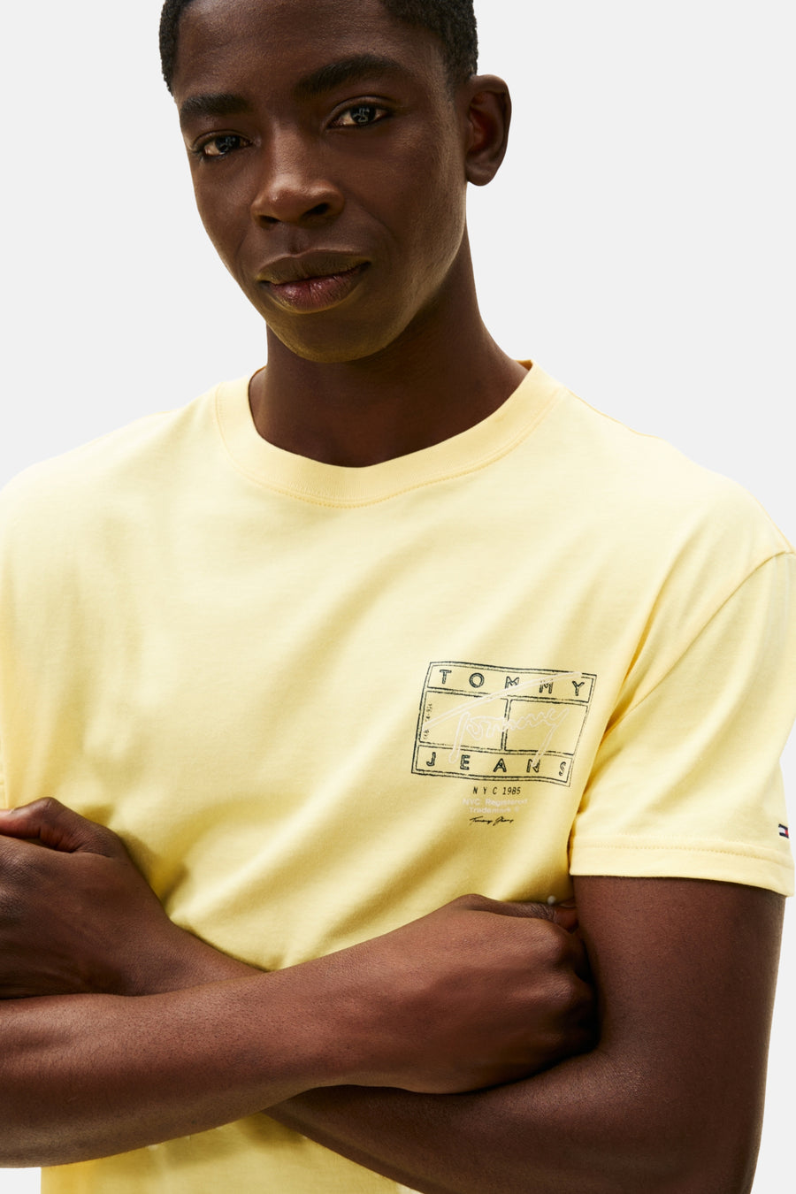 Een donkere man met gekruiste armen, geel Tommy Jeans T-shirt.
