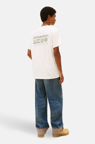Man van achteren in een wit T-shirt met groene "Tommy Jeans Bank Of" print, wijde blauwe jeans en gele schoenen, staand tegen een witte achtergrond.