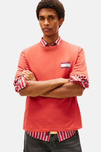 Man met gekruiste armen in rode Tommy Jeans-trui met gestreept overhemd eronder, tegen een witte achtergrond.