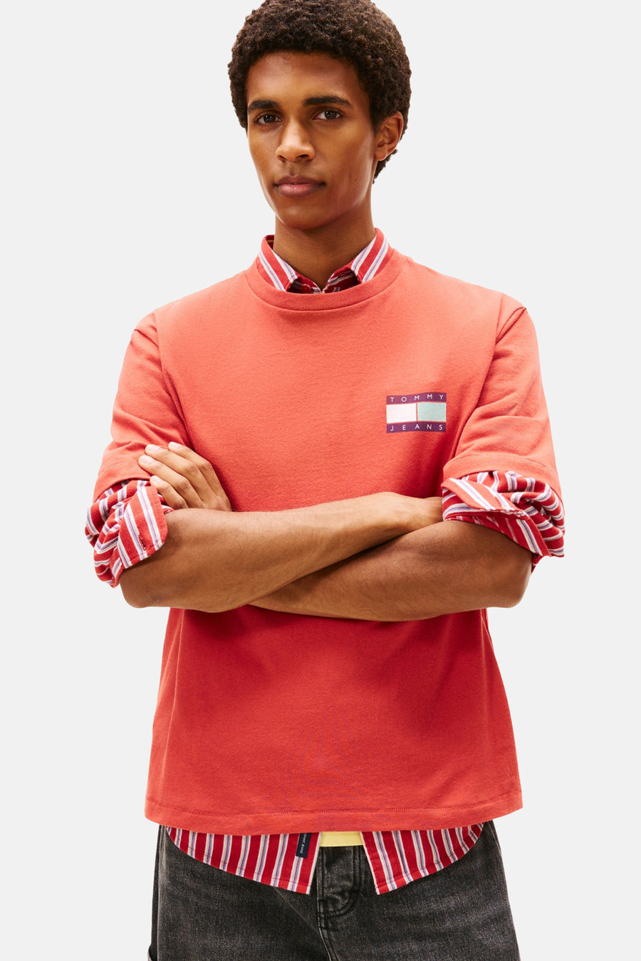 Man met gekruiste armen in rode Tommy Jeans-trui met gestreept overhemd eronder, tegen een witte achtergrond.