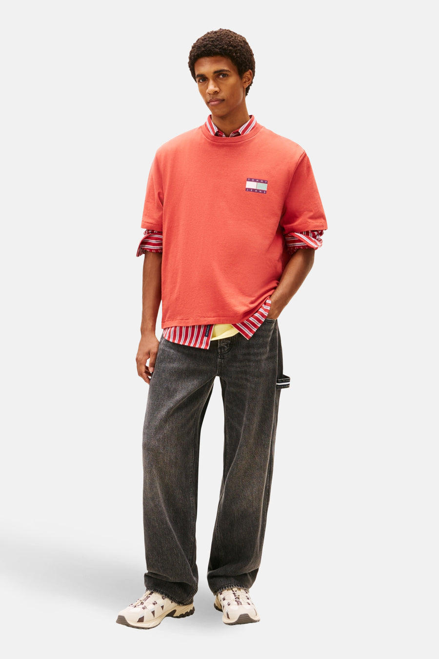 Man in rode Tommy Jeans T-shirt, gestreepte overhemd, zwarte jeans, beige sneakers, witte achtergrond.
