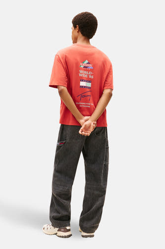 T-shirt rouge à manches courtes de TOMMY JEANS, avec une impression de logo sur le dos et associé à des jeans gris foncé et des chaussures crème.