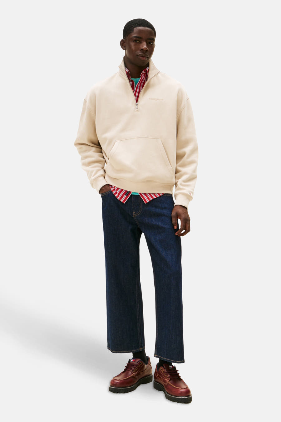 Gilet beige de TOMMY JEANS, avec une fermeture à glissière sur la moitié et un col montant, porté sur une chemise rayée rouge.