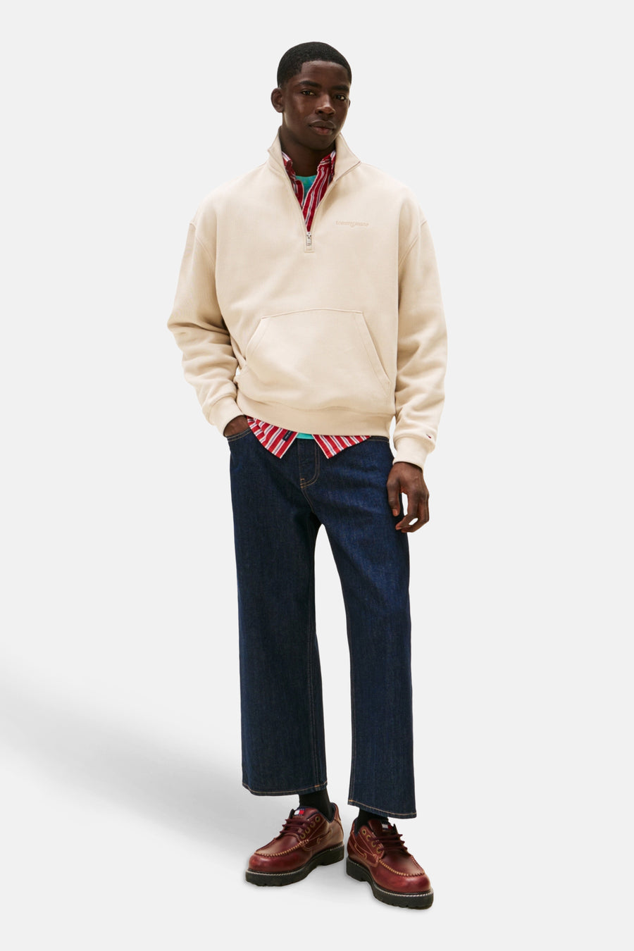 Man staand, beige trui, blauwe jeans, rode schoenen.