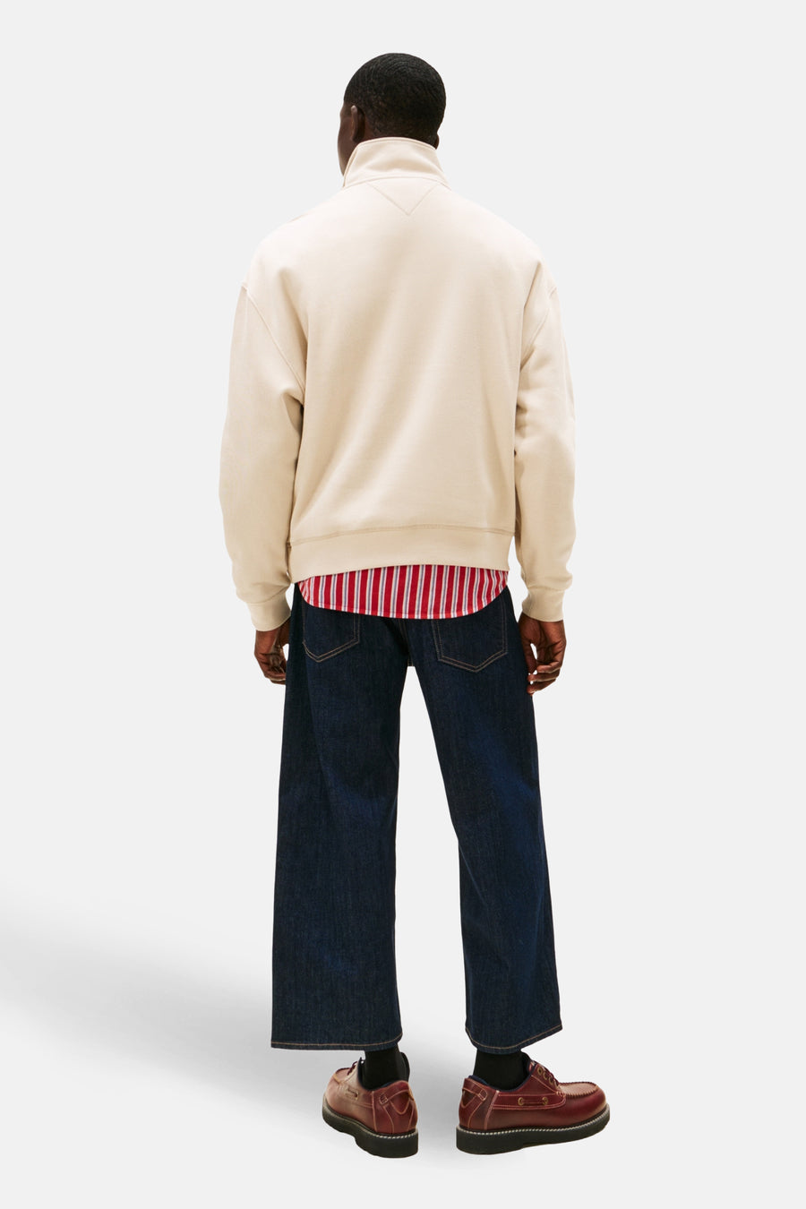 Man van achteren in jeans, beige trui, gestreept overhemd, en bruine schoenen, tegen een witte achtergrond.