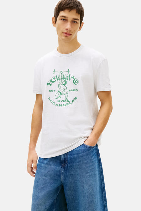T-shirt gris à manches courtes Tommy Jeans, avec imprimé baskets vert, combiné avec un pantacourt en jean et un pull vert sur les épaules.