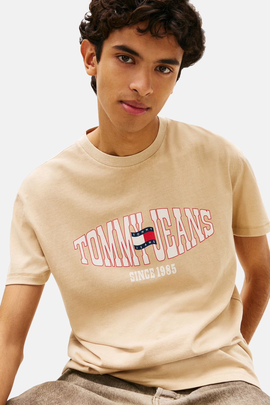 T-shirt beige de TOMMY JEANS avec le texte incurvé "TOMMY JEANS", un logo drapeau et "SINCE 1985" en dessous, porté par un mannequin.