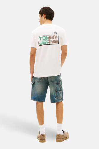 T-shirt écru à manches courtes de TOMMY JEANS, avec un imprimé graphique au dos, associé à un short en jean bleu, des chaussettes blanches et des chaussures en daim.
