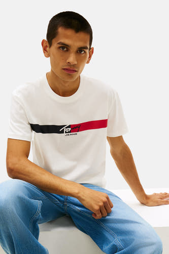 T-shirt blanc à manches courtes de TOMMY JEANS, avec une rayure rouge et noire sur la poitrine, combiné avec un jean bleu.