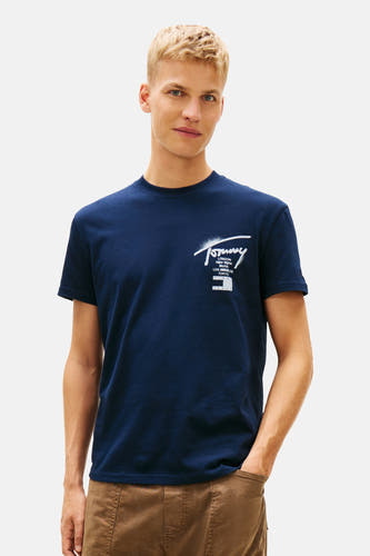T-shirt bleu à manches courtes de TOMMY JEANS, avec un pull sur les épaules et un pantalon marron.