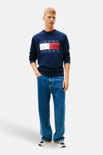 Pull-over bleu de TOMMY JEANS, avec un logo rectangulaire sur la poitrine, associé à des jeans et des baskets.