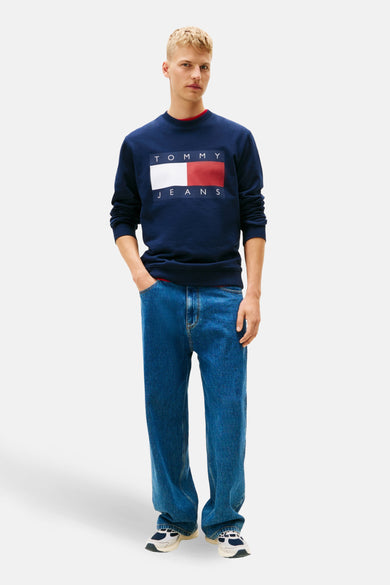 Pull-over bleu de TOMMY JEANS, avec un logo rectangulaire sur la poitrine, associé à des jeans et des baskets.