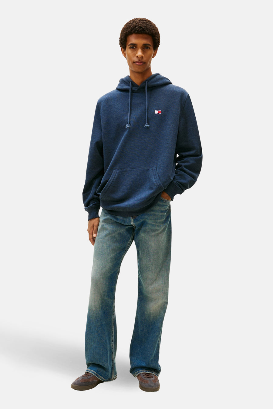 Man met donkerblauwe hoodie, jeans en bruine schoenen staand voor een witte achtergrond.
