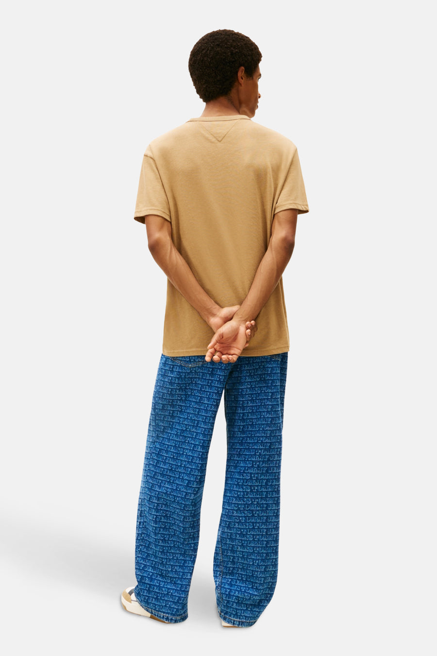 Achterkant van een man in bruin T-shirt en blauwe jeans met logo-print, staand op een witte achtergrond.