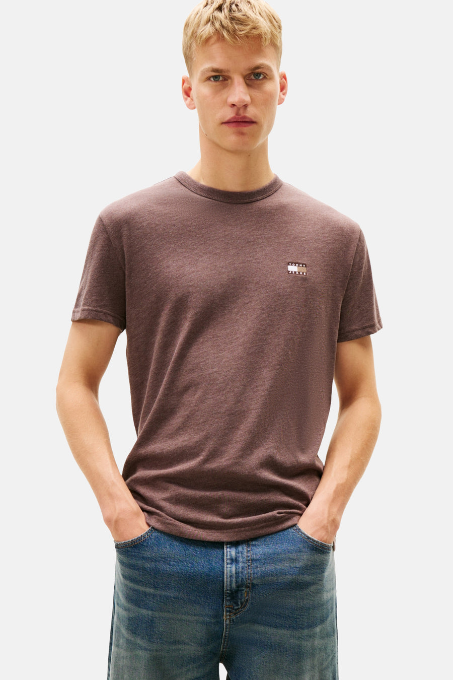 Man met bruin T-shirt en blauwe jeans, handen in zakken.
