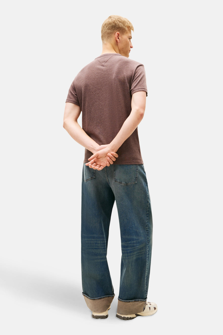 Achterkant van een man met bruin T-shirt en wijde spijkerbroek, handen gekruist achter de rug.