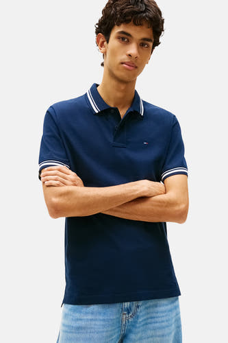 Polo bleu à manches courtes de TOMMY JEANS, avec des rayures blanches sur le col et les manches, combinée avec un jean ample et des mocassins noirs.