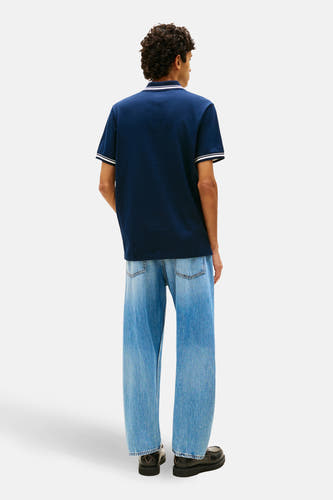 Polo bleu à manches courtes de TOMMY JEANS, avec des accents blancs sur le col et les manches. Porté avec un jean ample et des chaussures noires.