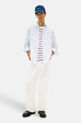 Blauw overhemd van TOMMY JEANS, gedragen over een gestreepte top, met witte broek en een blauwe pet.
