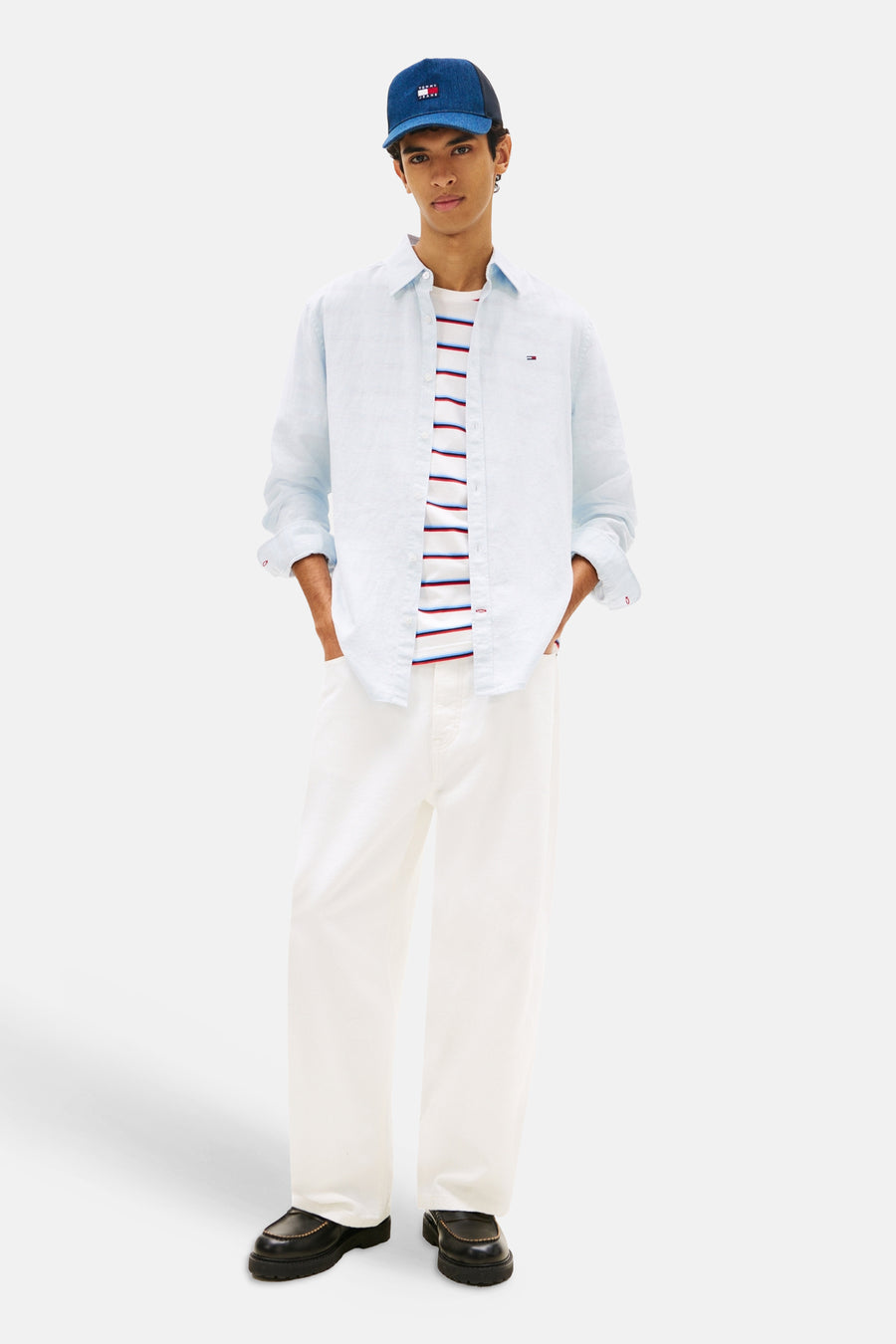 Blauw overhemd van TOMMY JEANS, gedragen over een gestreepte top, met witte broek en een blauwe pet.
