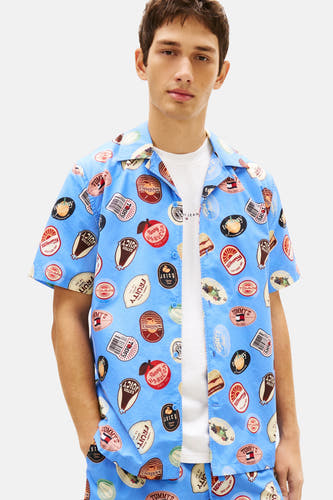 Chemise bleue à manches courtes Tommy Jeans, ornée d'un motif d'étiquettes répétées, portée avec un t-shirt blanc, un short et des baskets.