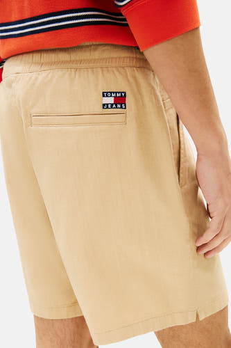 Short marron clair de TOMMY JEANS avec taille élastique, une poche passepoilée et un petit écusson de marque au dos. Porté par un homme avec une chemise à rayures rouges.