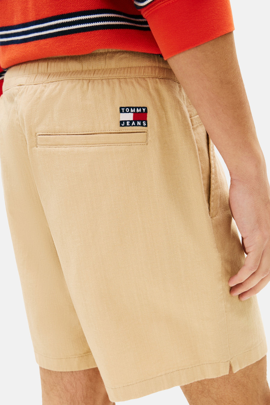 Short marron clair de TOMMY JEANS avec taille élastique, une poche passepoilée et un petit écusson de marque au dos. Porté par un homme avec une chemise à rayures rouges.