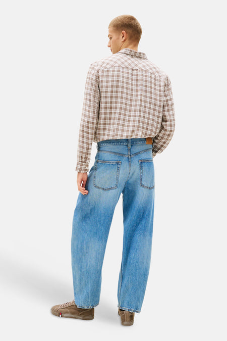 Jean en denim bleu clair à coupe ample de TOMMY JEANS, vu de dos sur un mannequin portant une chemise à carreaux et des baskets marron.