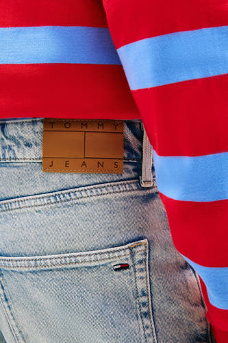 Short en jean denim bleu clair de TOMMY JEANS avec patch en cuir marron et petit logo drapeau, porté avec un pull à rayures rouge et bleu.
