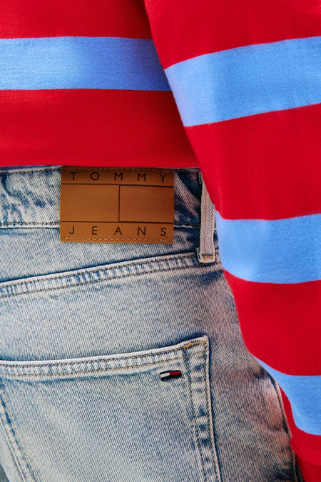 Short en jean denim bleu clair de TOMMY JEANS avec patch en cuir marron et petit logo drapeau, porté avec un pull à rayures rouge et bleu.