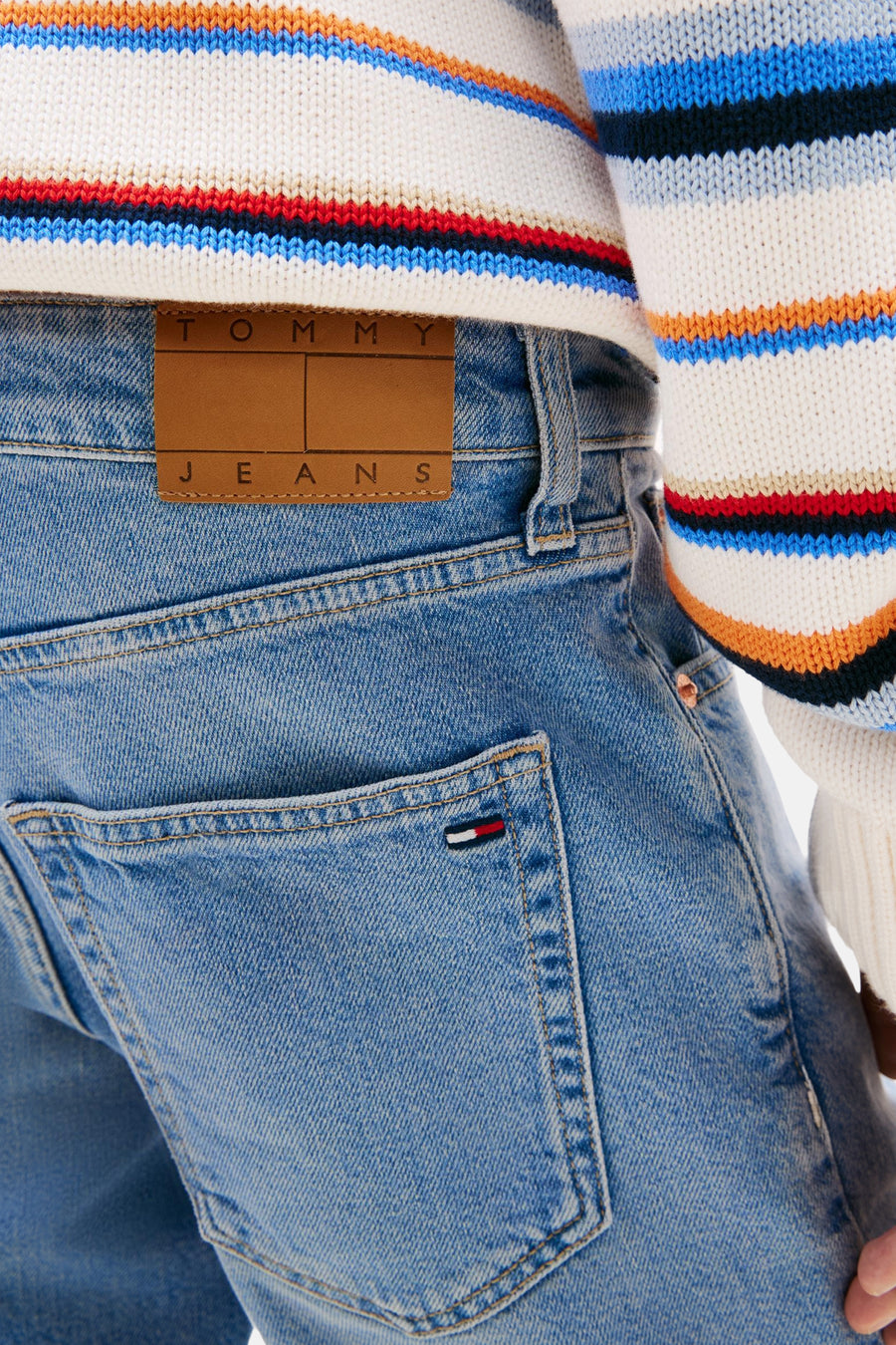 Short en jean bleu denim de TOMMY JEANS, avec patch logo en cuir et détail de drapeau sur la poche arrière, porté avec un pull rayé.