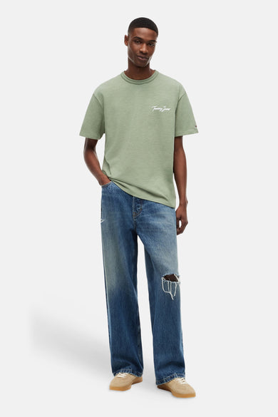 T-shirt vert à manches courtes TOMMY JEANS avec logo script blanc sur la poitrine, porté par un homme avec un jean déchiré et des baskets beiges.