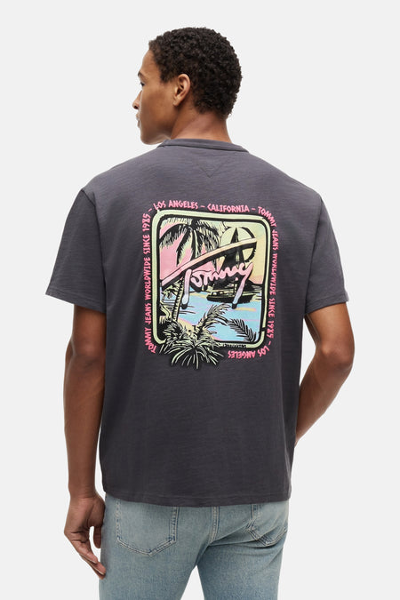 T-shirt gris à manches courtes TOMMY JEANS, imprimé plage rétro coloré et texte au dos, porté avec un jean.