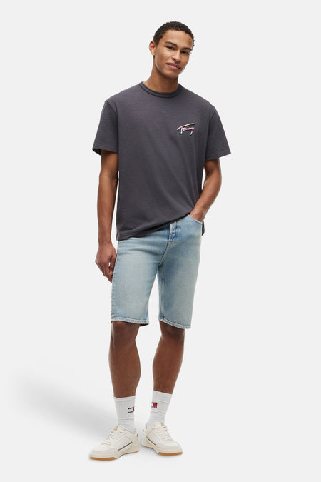 T-shirt gris à manches courtes de TOMMY JEANS, avec logo Tommy coloré, porté avec un short en jean bleu clair et des baskets de couleur crème.