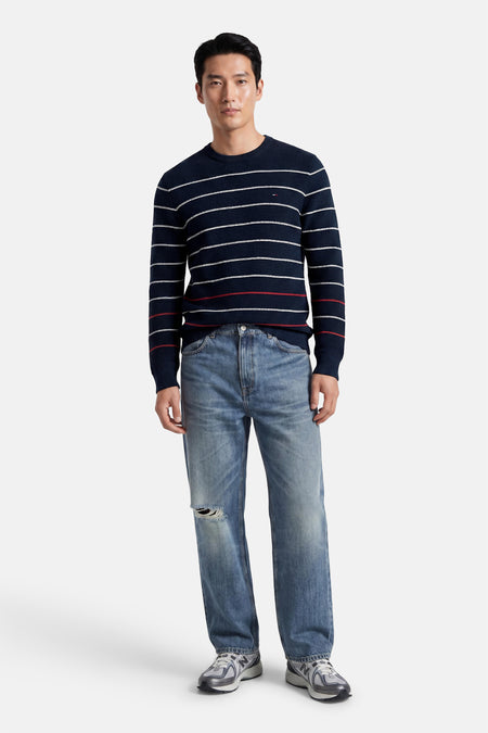 Blauwe ronde hals trui met witte en rode strepen van TOMMY JEANS, gedragen met grijze jeans en sneakers, front view