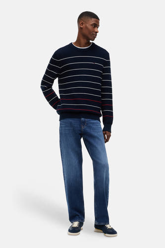Pull bleu à col rond de TOMMY JEANS, avec des rayures blanches et rouges, en maille côtelée, porté par un homme avec un jean et des baskets.