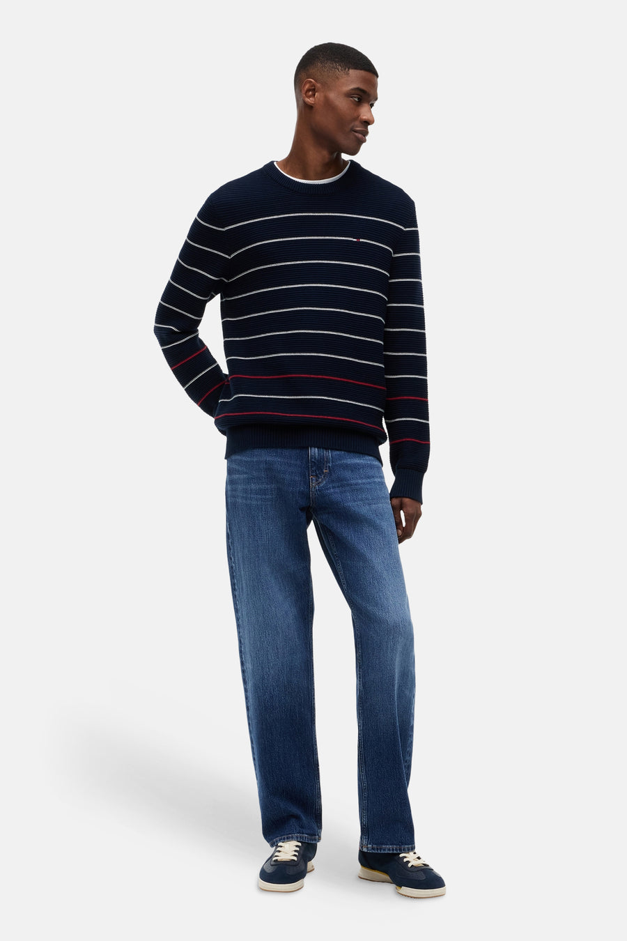 Pull bleu à col rond de TOMMY JEANS, avec des rayures blanches et rouges, en maille côtelée, porté par un homme avec un jean et des baskets.