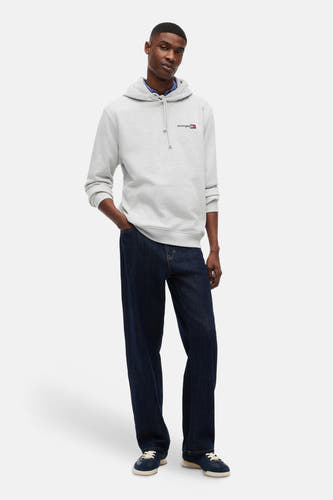 Sweat-shirt gris TOMMY JEANS à capuche avec logo sur la poitrine, porté sur une chemise bleue avec un jean bleu foncé et des baskets bleues.