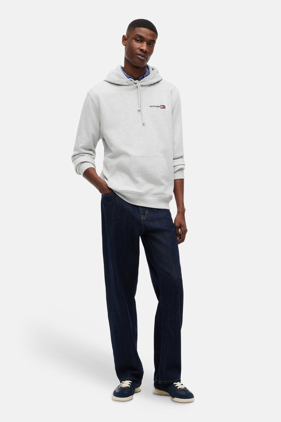 Sweat-shirt gris TOMMY JEANS à capuche avec logo sur la poitrine, porté sur une chemise bleue avec un jean bleu foncé et des baskets bleues.