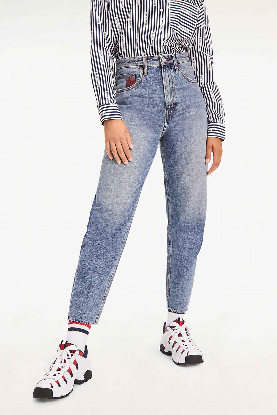 Femme en tenue Tommy Jeans, chemise rayée, jeans bleus et baskets blanches avec des accents rouges.