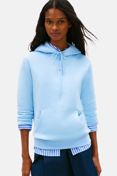 Sweatshirt à capuche bleu de TOMMY JEANS, avec cordon de serrage et poche kangourou, porté sur une chemise rayée et une minijupe plissée, associé à des chaussettes et des baskets.