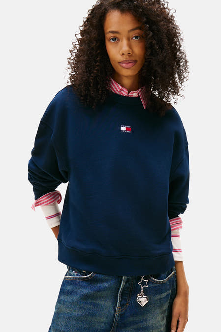 Pull-over bleu de TOMMY JEANS, porté avec un chemisier rayé et une pantalon en cuir avec un cordon.