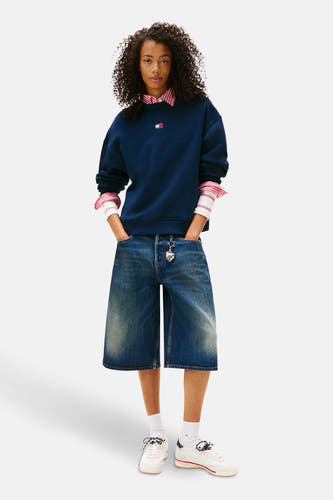 Pull-over bleu de TOMMY JEANS, porté sur un chemisier rayé, avec des bermudas en denim large et des baskets blanches.