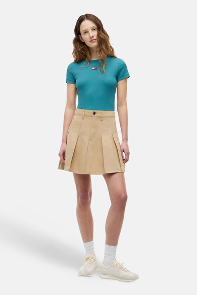 T-shirt côtelé vert essence de TOMMY JEANS avec petit logo et manches courtes, porté par un mannequin avec une minijupe beige et des baskets blanches.